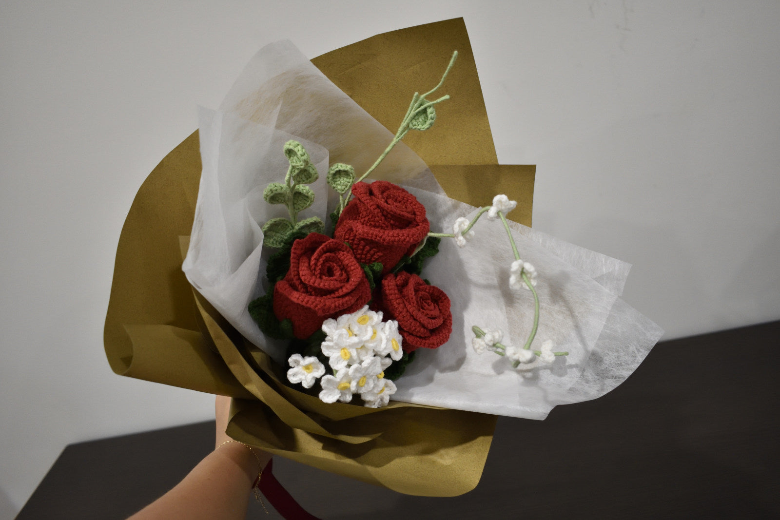 Handmade crochet flower bouquet featuring three dark red roses with white accent flowers and greenery, wrapped in brown paper and held against a neutral background.