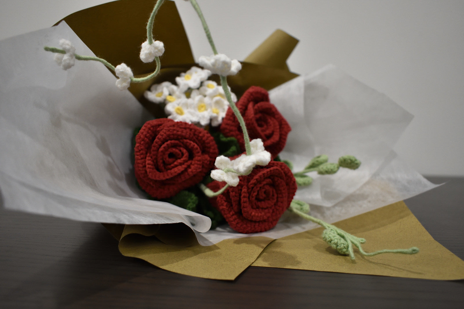 Handmade crochet flower bouquet with three dark red roses, white accent flowers, and greenery, wrapped in brown paper and laid on a dark table.