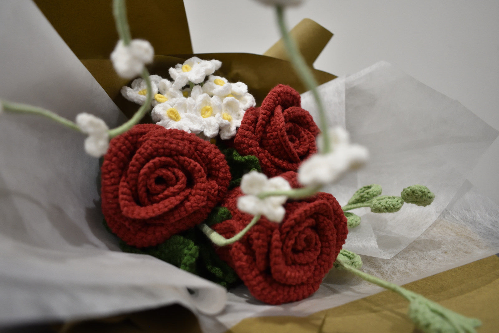 Close-up of a handmade crochet flower bouquet with three dark red roses, white accent flowers, and greenery, wrapped in brown paper.