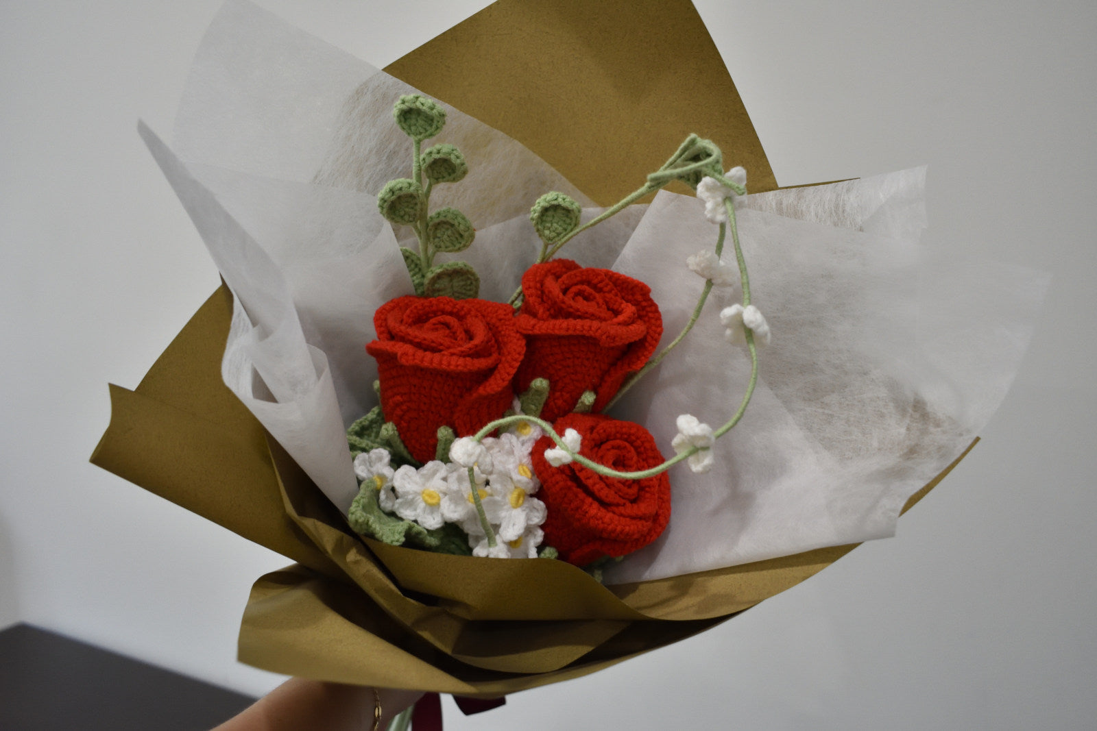 Handmade crochet flower bouquet featuring three red roses with white accent flowers and greenery, wrapped in brown paper and held against a neutral background.