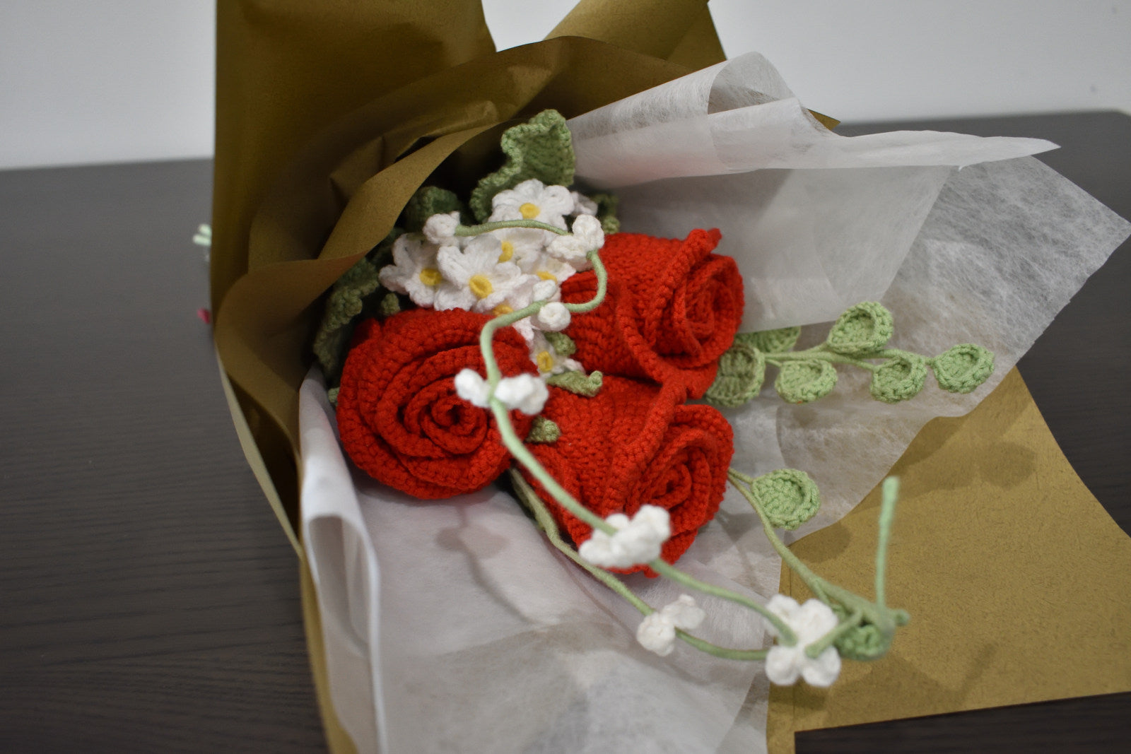 Handmade crochet flower bouquet featuring three red roses with white accent flowers and greenery, wrapped in brown paper and laid on a dark table.