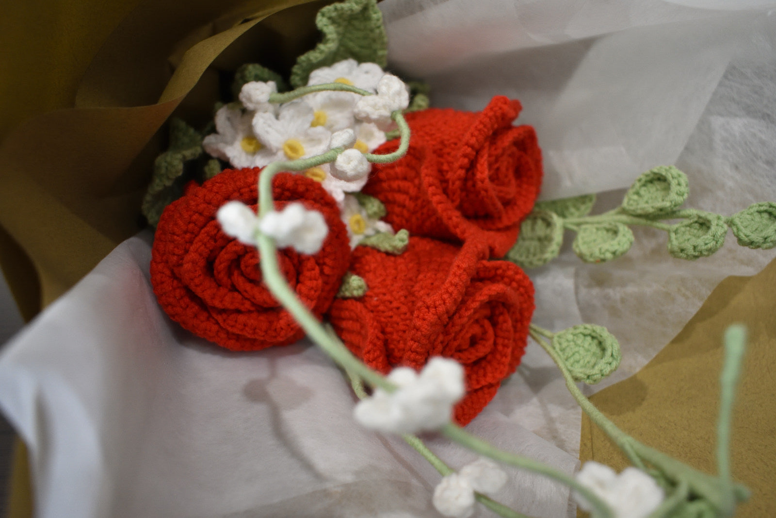 Close-up of a handmade crochet flower bouquet featuring three red roses with white accent flowers and greenery, wrapped in brown paper.