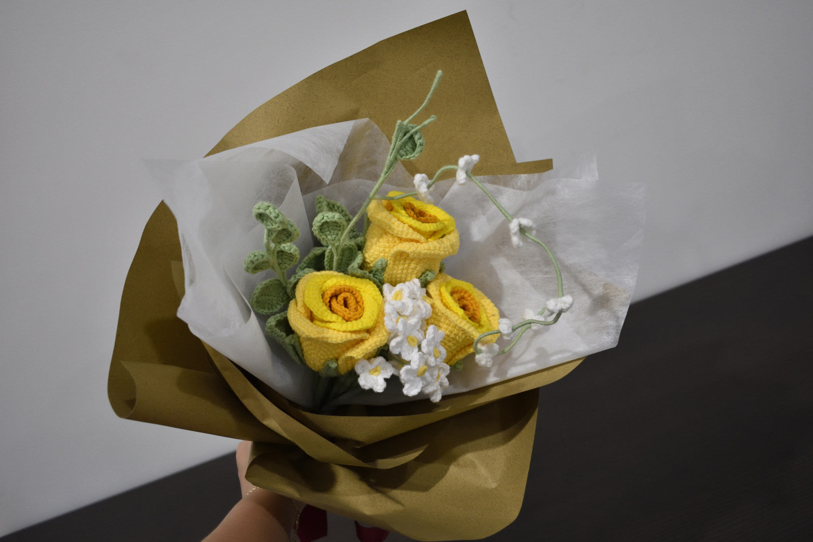 Handmade crochet flower bouquet featuring three yellow roses with white accent flowers and greenery, wrapped in brown paper and held against a neutral background.