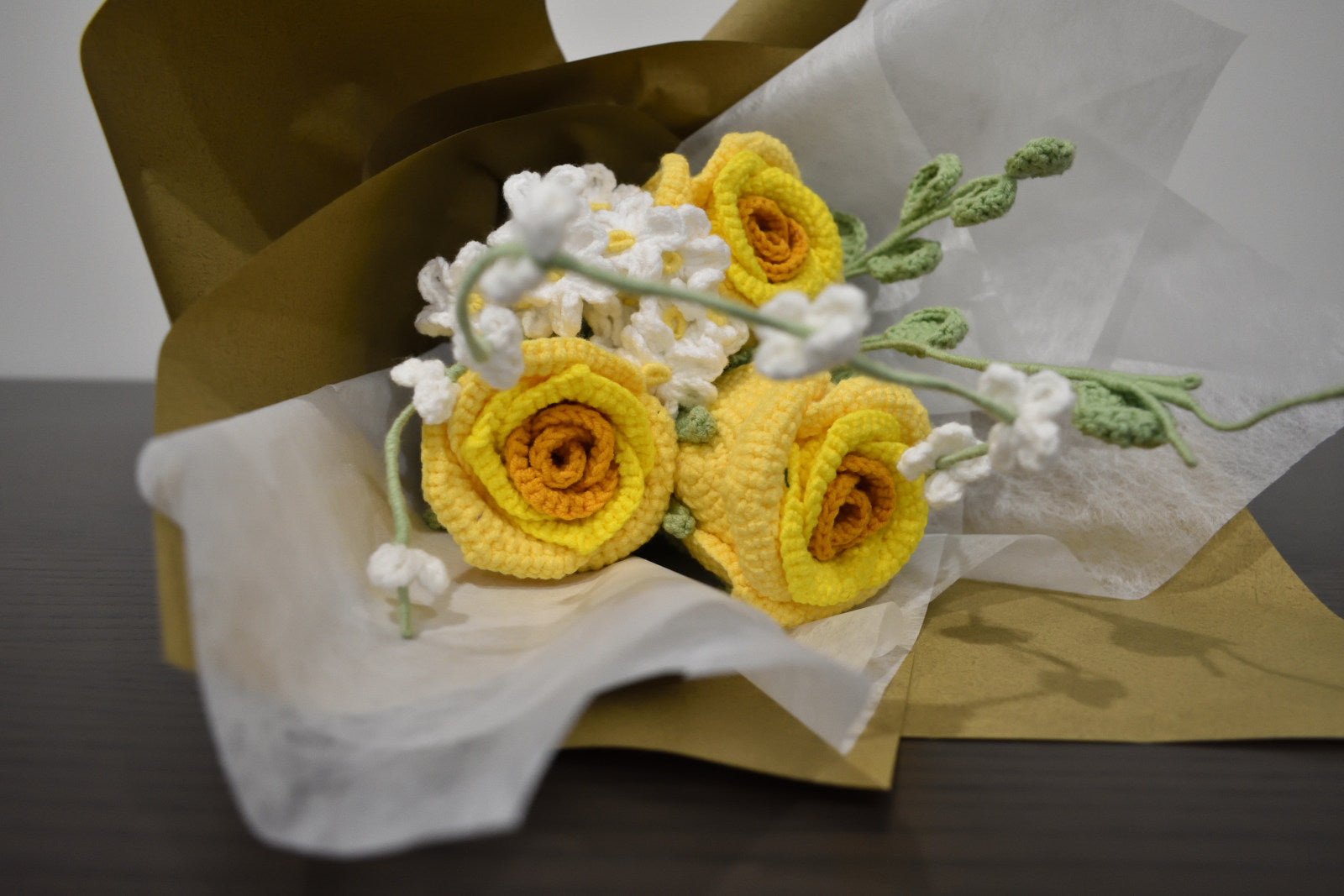Handmade crochet flower bouquet featuring three yellow roses with white accent flowers and greenery, wrapped in brown paper and laid on a dark table.