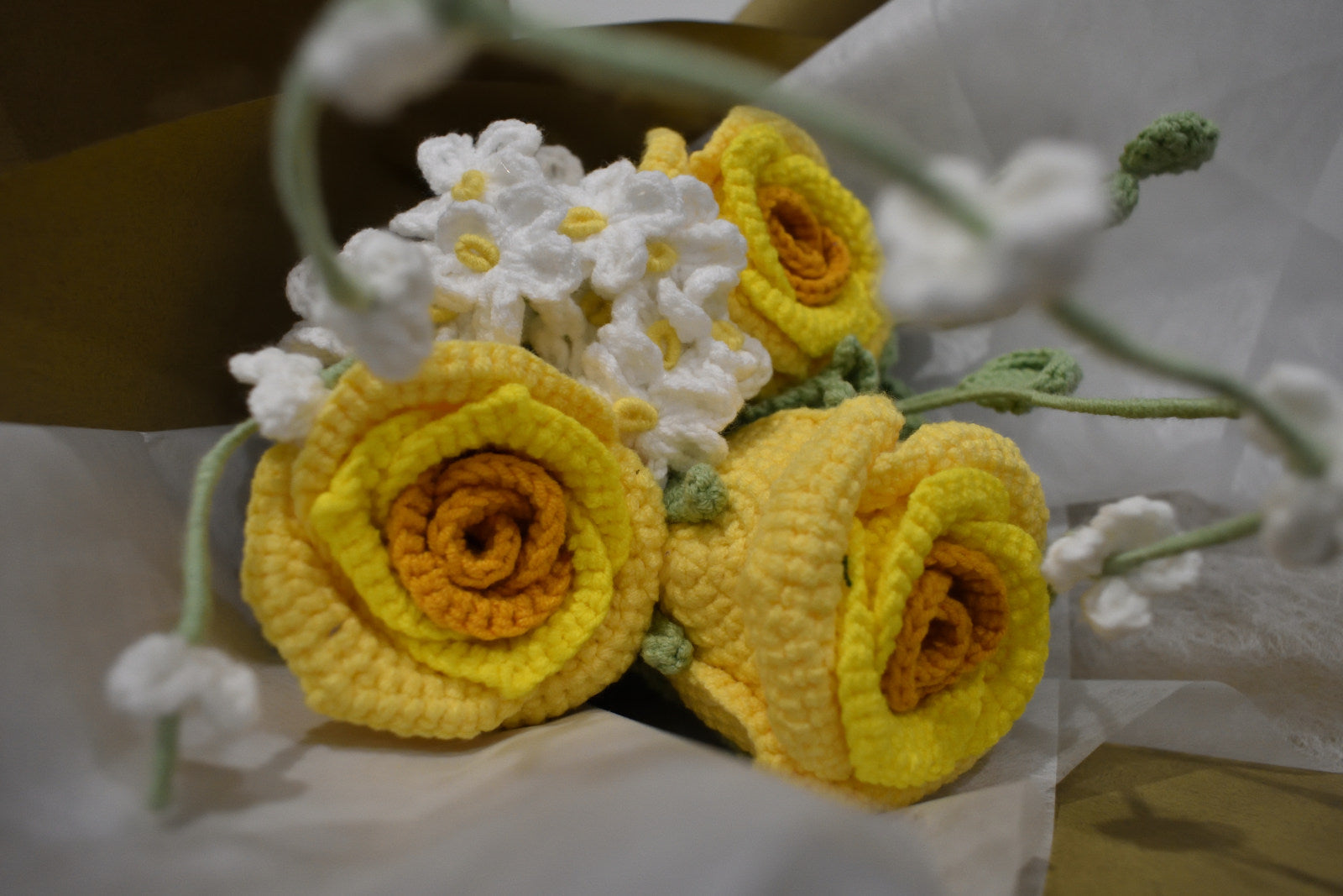 Close-up of a handmade crochet flower bouquet featuring three yellow roses with white accent flowers and greenery, wrapped in brown paper.