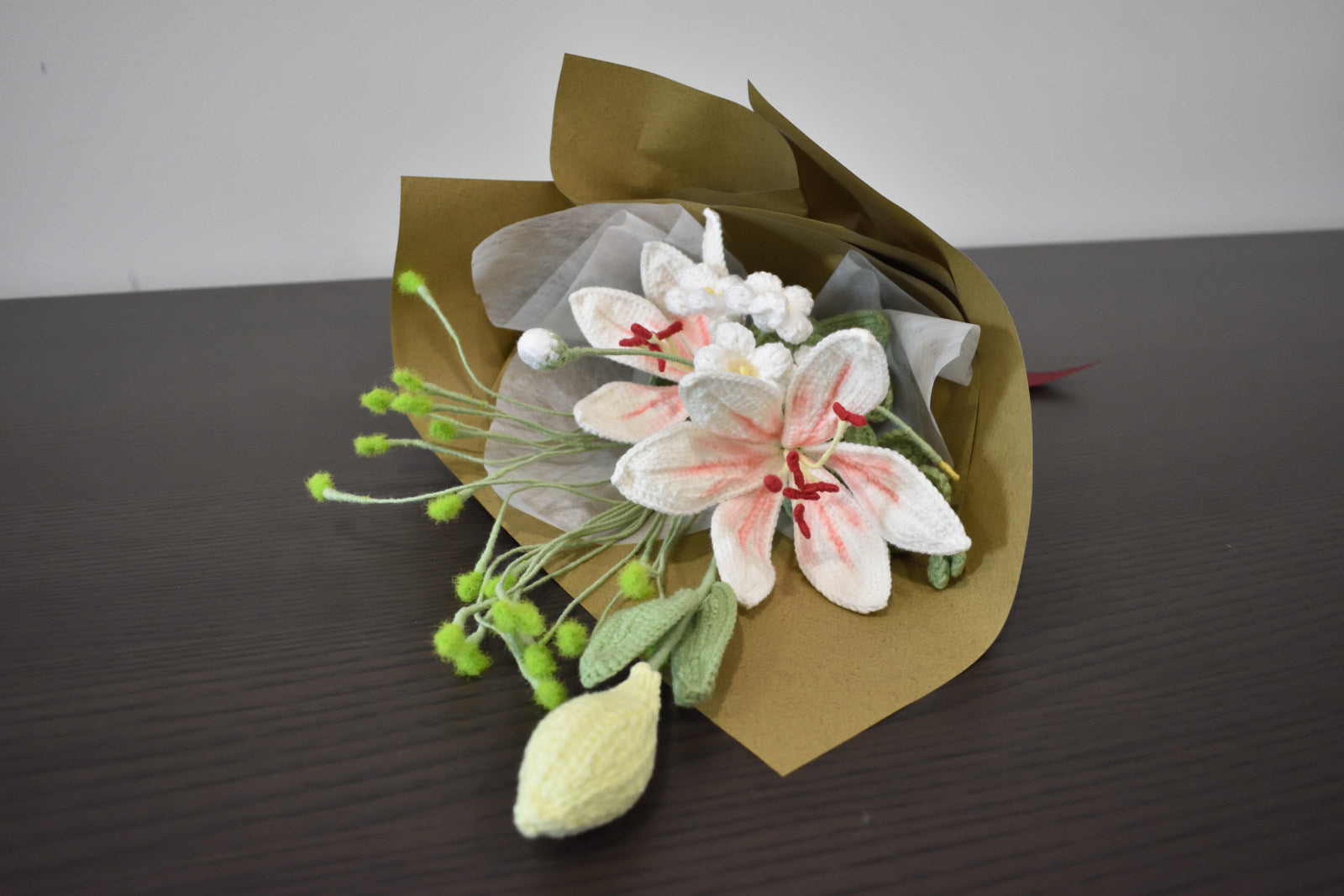 Handmade crochet flower bouquet featuring twin pink and white lilies with one flower bud, wrapped in brown paper and laid on a dark table.