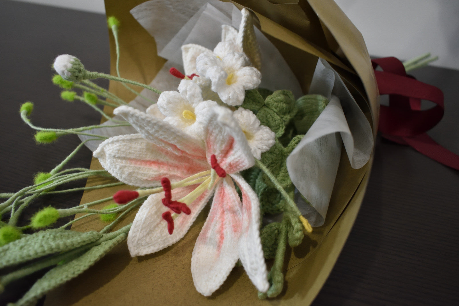 Close-up of a handmade crochet flower bouquet featuring twin pink and white lilies with one flower bud, wrapped in brown paper and laid on a dark table.