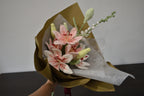 Handmade crochet flower bouquet featuring three pink lilies, two white and pink lilies, an oversized delphinium, and a snowberry branch, wrapped in brown paper and held against a neutral background.