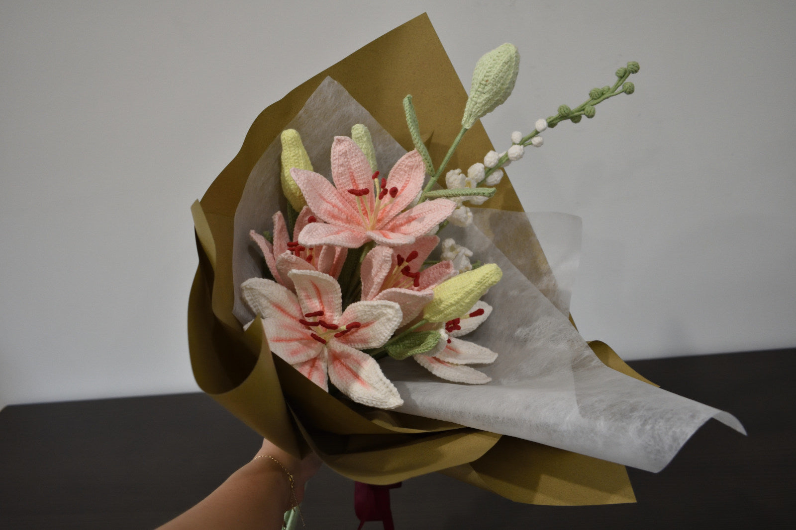Handmade crochet flower bouquet featuring three pink lilies, two white and pink lilies, an oversized delphinium, and a snowberry branch, wrapped in brown paper and held against a neutral background.