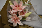 Close-up of a handmade crochet flower bouquet featuring three pink lilies, two white and pink lilies, an oversized delphinium, and a snowberry branch, wrapped in brown paper.