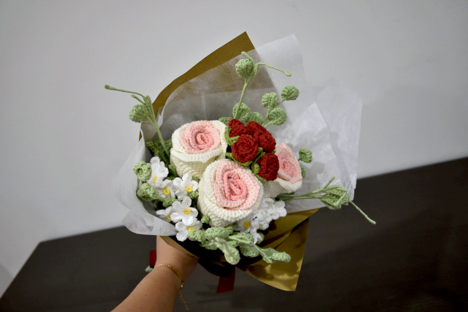 Handmade crochet flower bouquet in pink and white with red accent roses and greenery, wrapped in brown paper and held against a neutral background.