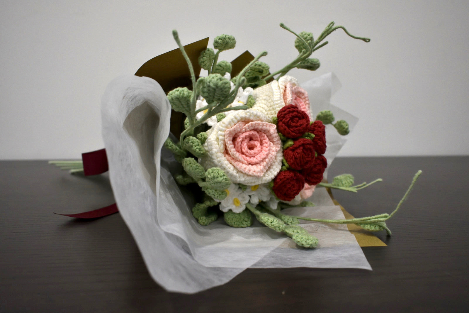 Handmade crochet flower bouquet in pink and white with red accent roses and greenery, wrapped in brown paper and laid on a dark table.