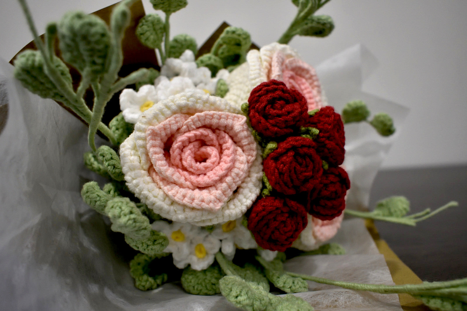 Close-up of a handmade crochet flower bouquet in pink and white with red accent roses and greenery, wrapped in brown paper and laid on a dark table.