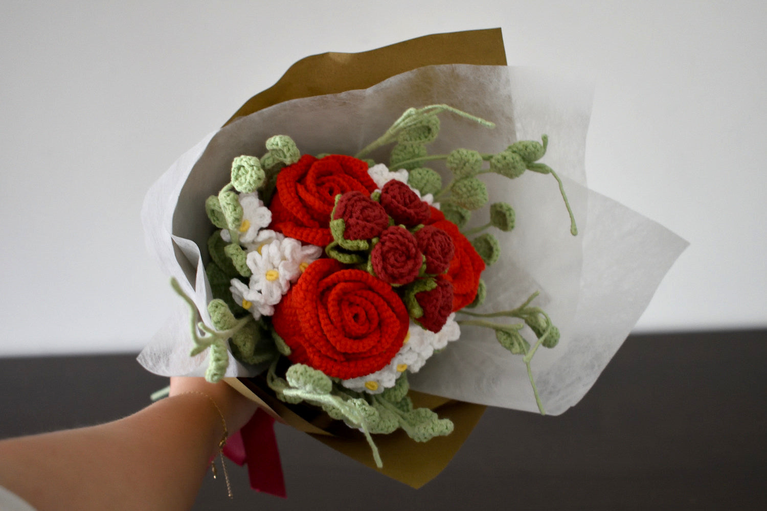 Handmade crochet flower bouquet in red with red accent roses and greenery, wrapped in brown paper and held against a neutral background.