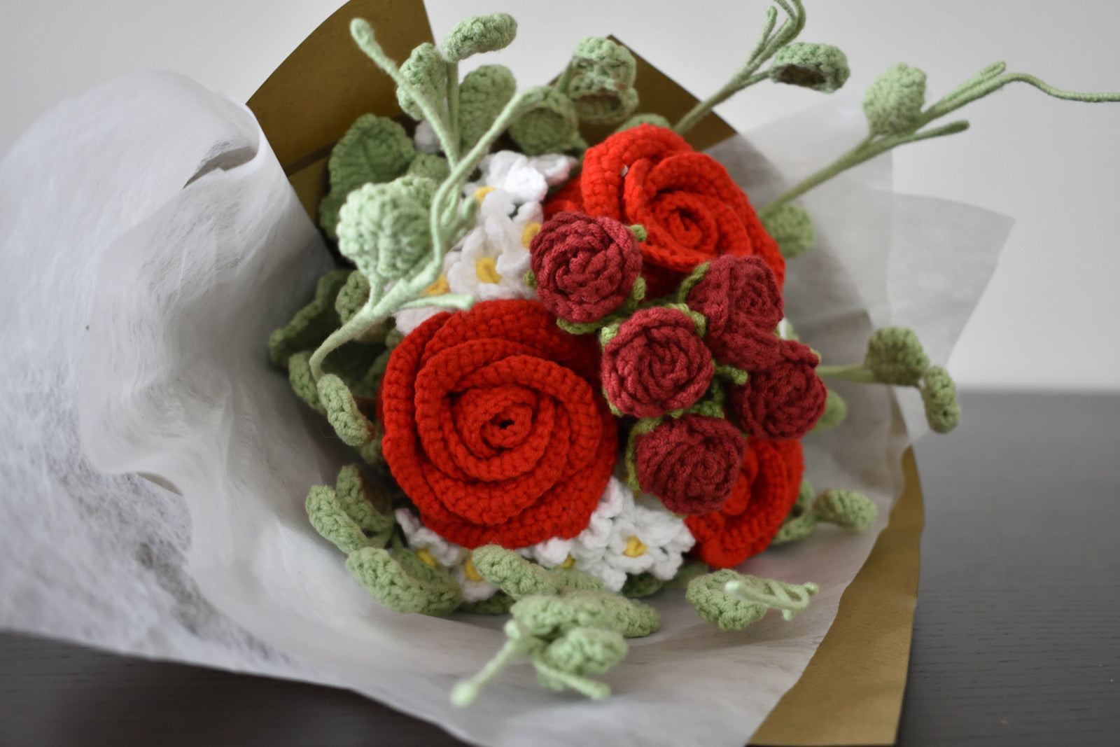 Handmade crochet flower bouquet in red with red accent roses and greenery, wrapped in brown paper and laid on a dark table.