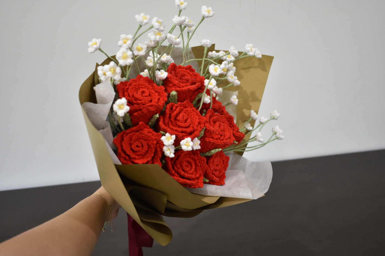 Handmade crochet flower bouquet featuring nine red roses with baby's breath, wrapped in brown paper and held against a neutral background.