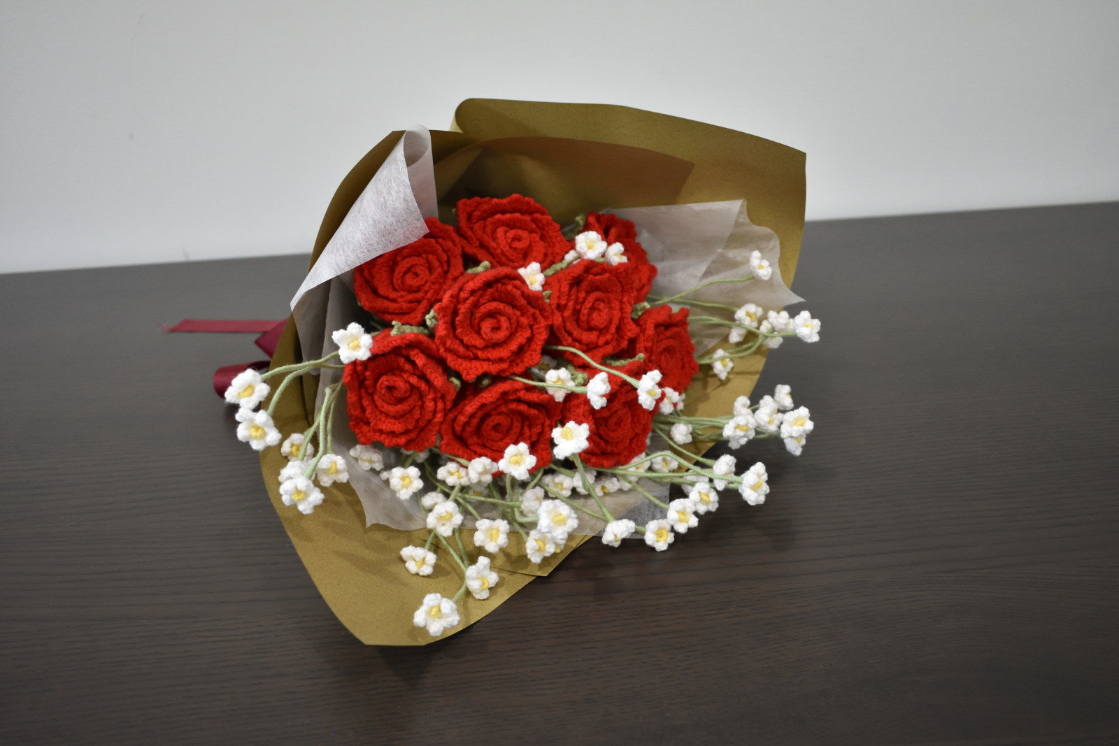 Handmade crochet flower bouquet featuring nine red roses with baby's breath, wrapped in brown paper and laid on a dark table.