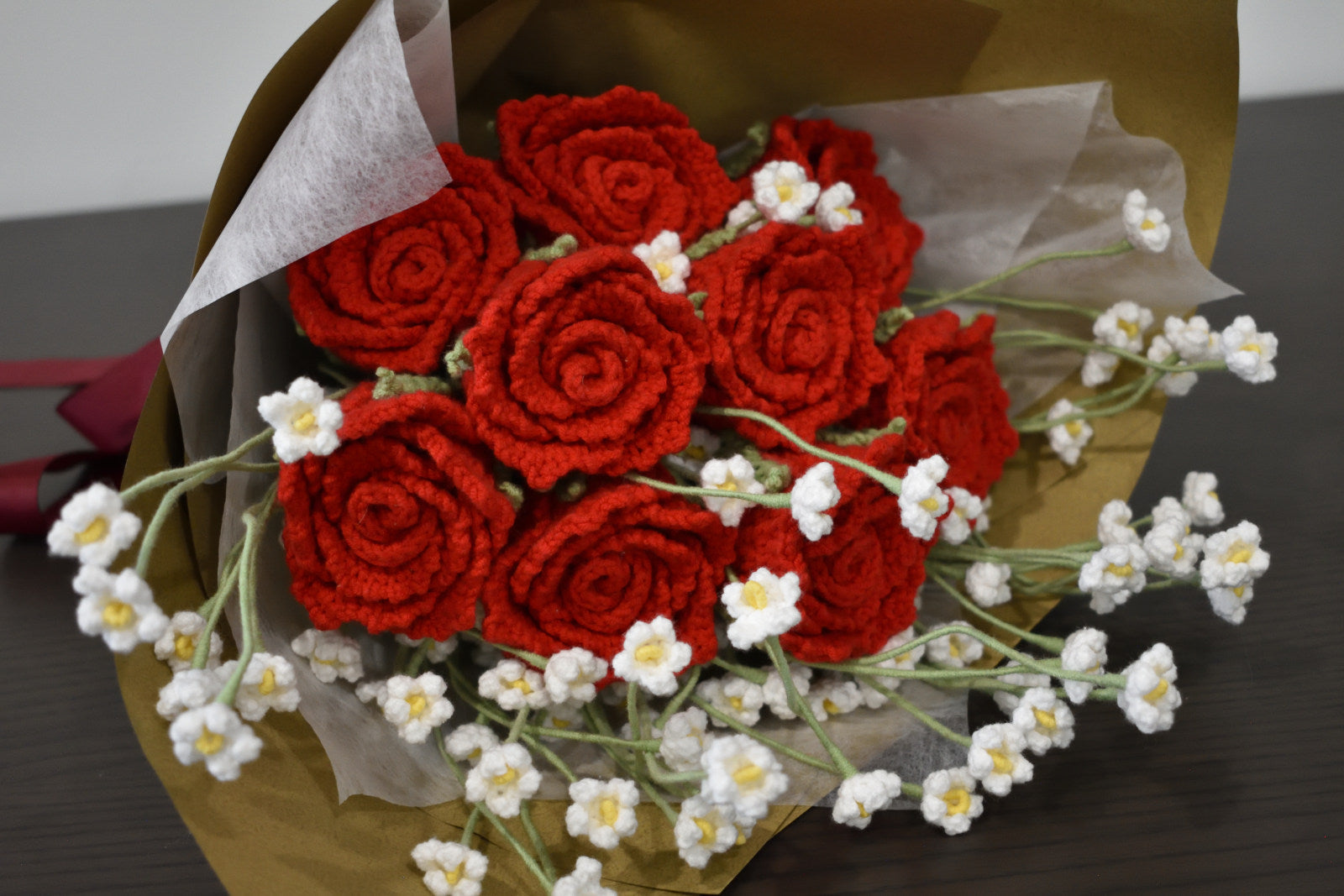 Close-up of a handmade crochet flower bouquet featuring nine red roses with baby's breath, wrapped in brown paper and laid on a dark table.