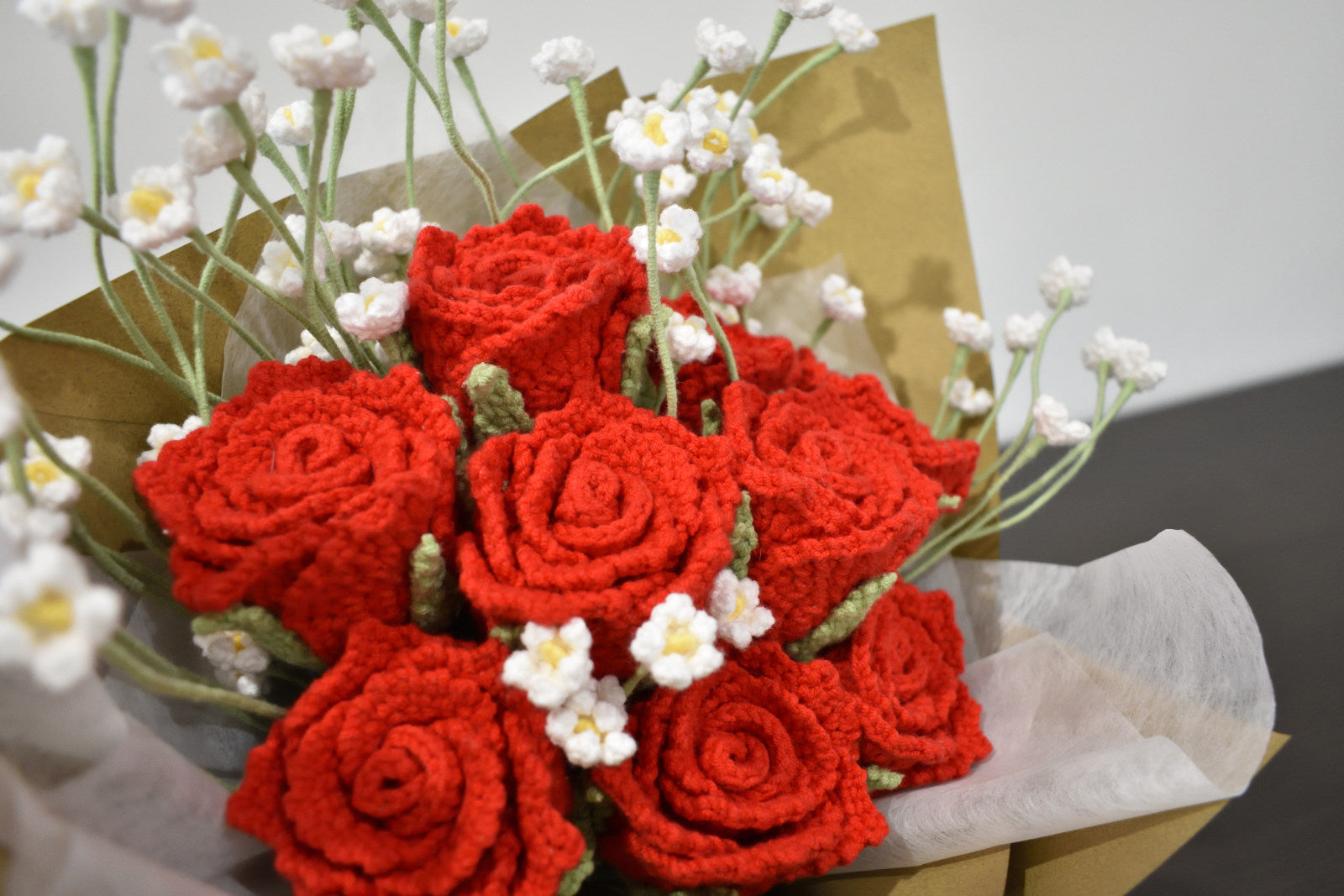 Close-up of a handmade crochet flower bouquet featuring nine red roses with baby's breath, wrapped in brown paper and held against a neutral background.