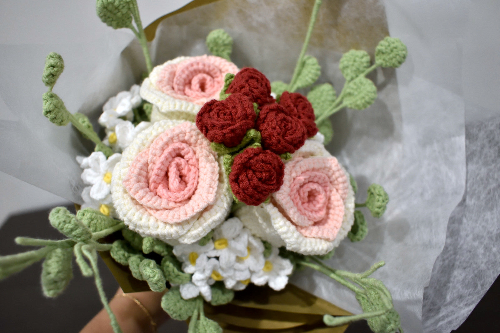 Close-up of a handmade crochet flower bouquet in pink and white with red accent roses and greenery, wrapped in brown paper, held against a neutral background.