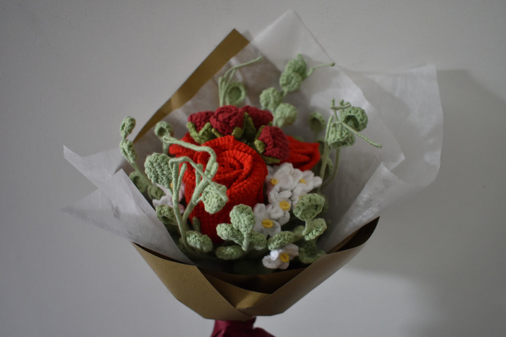 Close up of a handmade crochet flower bouquet in pink and white with red accent roses and greenery, wrapped in brown paper and held against a neutral background.