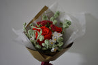 Close up of a handmade crochet flower bouquet in pink and white with red accent roses and greenery, wrapped in brown paper and held against a neutral background.