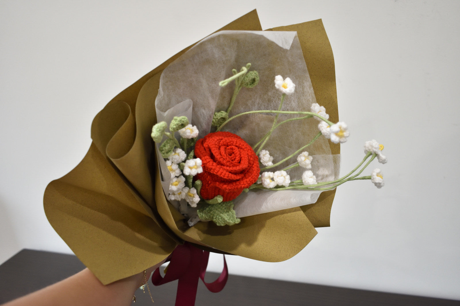 Handmade crochet flower rose bouquet featuring a single red rose with baby's breath and greenery, wrapped in brown paper and held against a neutral background.
