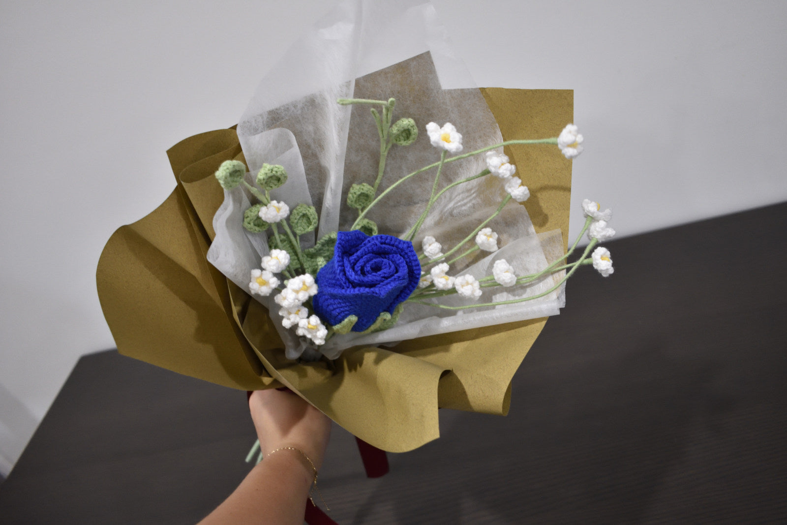 Handmade crochet flower bouquet featuring a single blue rose with baby's breath and greenery, wrapped in brown paper and held against a neutral background.