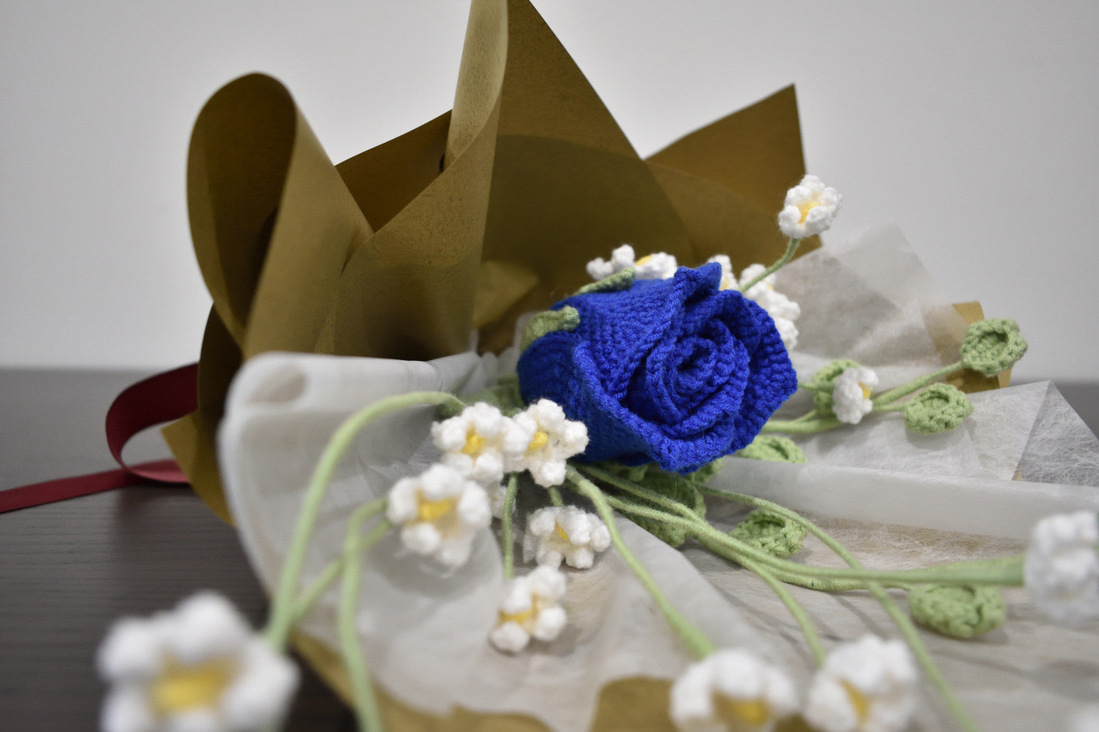 Handmade crochet flower bouquet featuring a single blue rose with baby's breath and greenery, wrapped in brown paper and laid on a dark table.