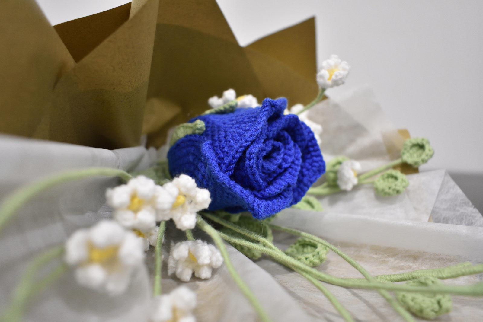 Close-up of a handmade crochet flower bouquet featuring a single blue rose with baby's breath and greenery, wrapped in brown paper and laid on a dark table.