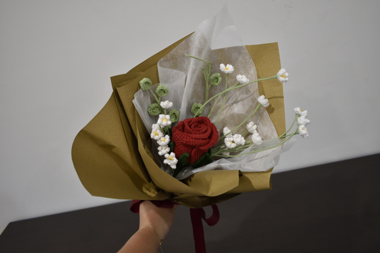 Handmade crochet flower bouquet featuring a single dark red rose with baby's breath and greenery, wrapped in brown paper and held against a neutral background.