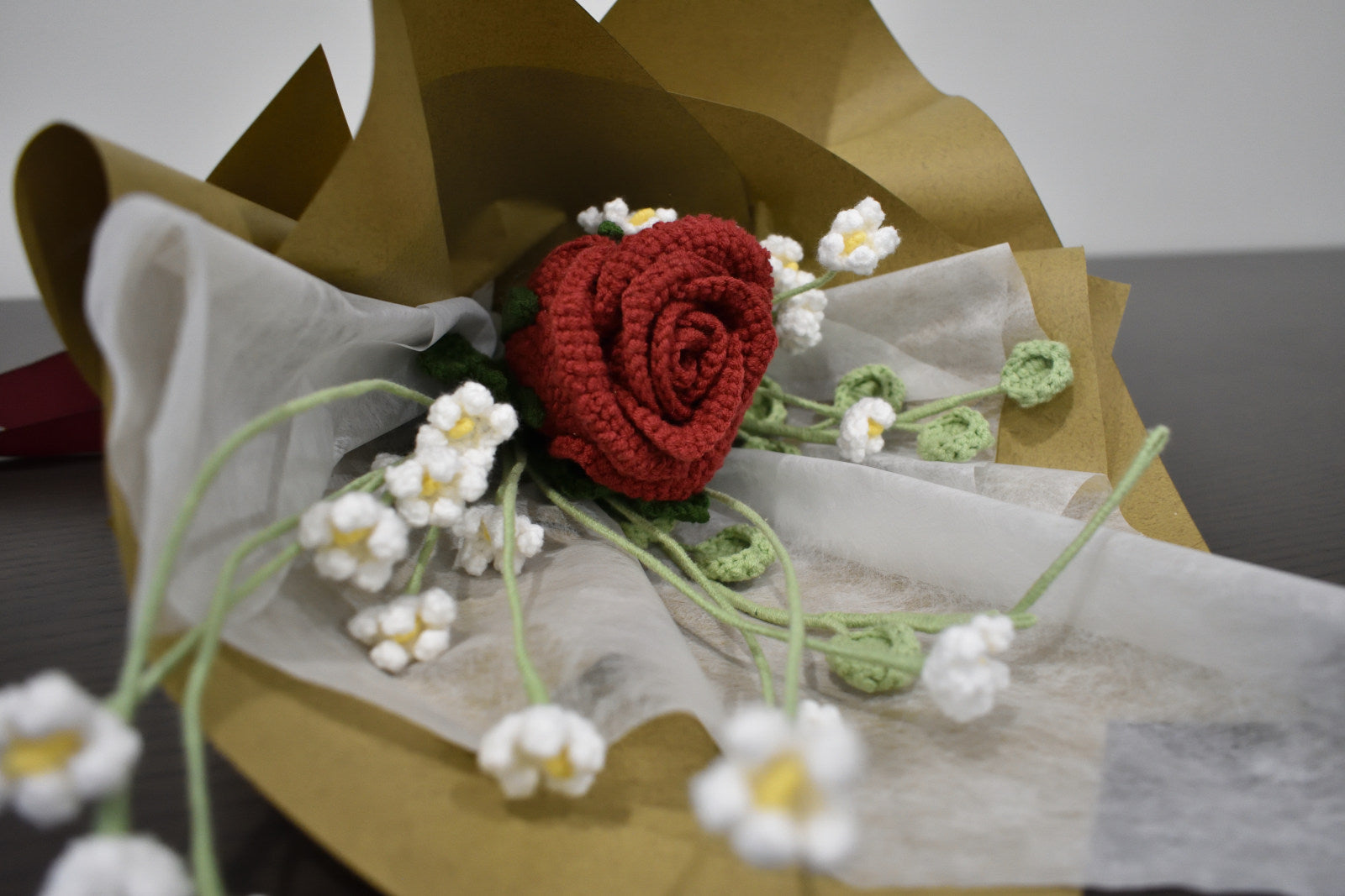 Handmade crochet flower bouquet featuring a single dark red rose with baby's breath and greenery, wrapped in brown paper and laid on a dark table.