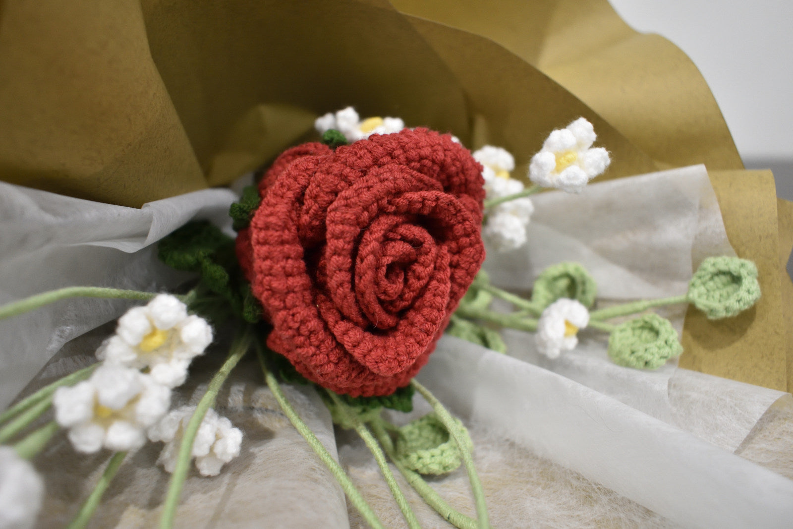 Close-up of a handmade crochet flower bouquet featuring a single dark red rose with baby's breath and greenery, wrapped in brown paper.