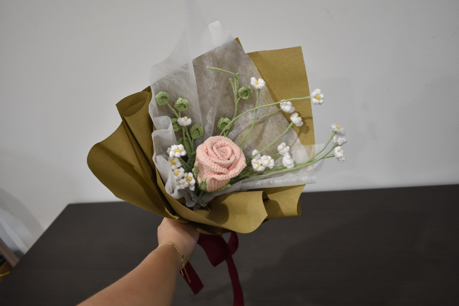 Handmade crochet flower bouquet featuring a single pink rose with baby's breath and greenery, wrapped in brown paper and held against a neutral background.