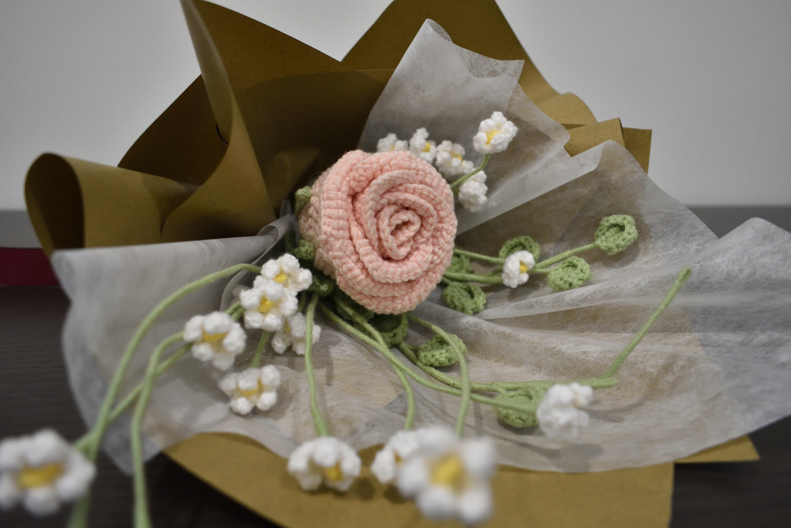 Handmade crochet flower bouquet featuring a single pink rose with baby's breath and greenery, wrapped in brown paper and laid on a dark table.