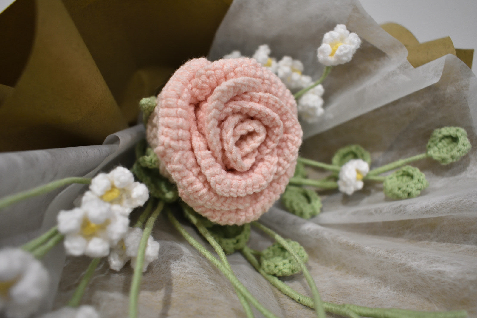 Close-up of a handmade crochet flower bouquet featuring a single pink rose with baby's breath and greenery, wrapped in brown paper and laid on a dark table.
