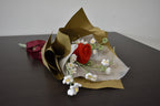 Handmade crochet flower bouquet featuring a single red rose with baby's breath and greenery, wrapped in brown paper and laid on a dark table.