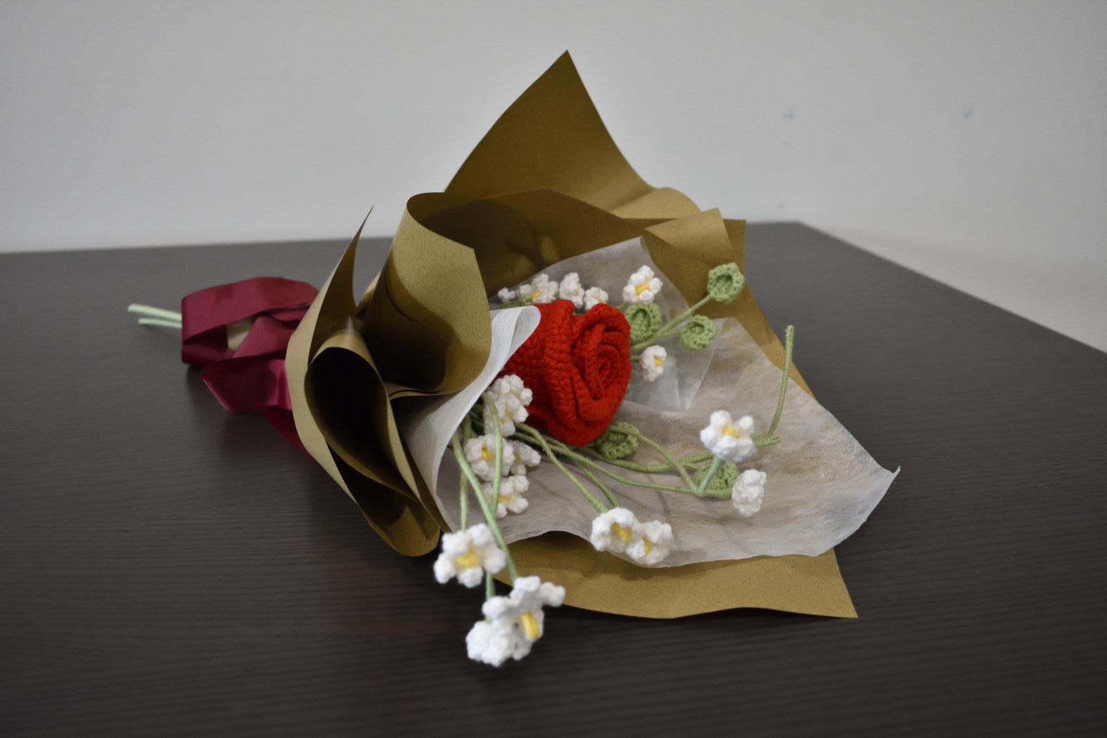 Handmade crochet flower bouquet featuring a single red rose with baby's breath and greenery, wrapped in brown paper and laid on a dark table.