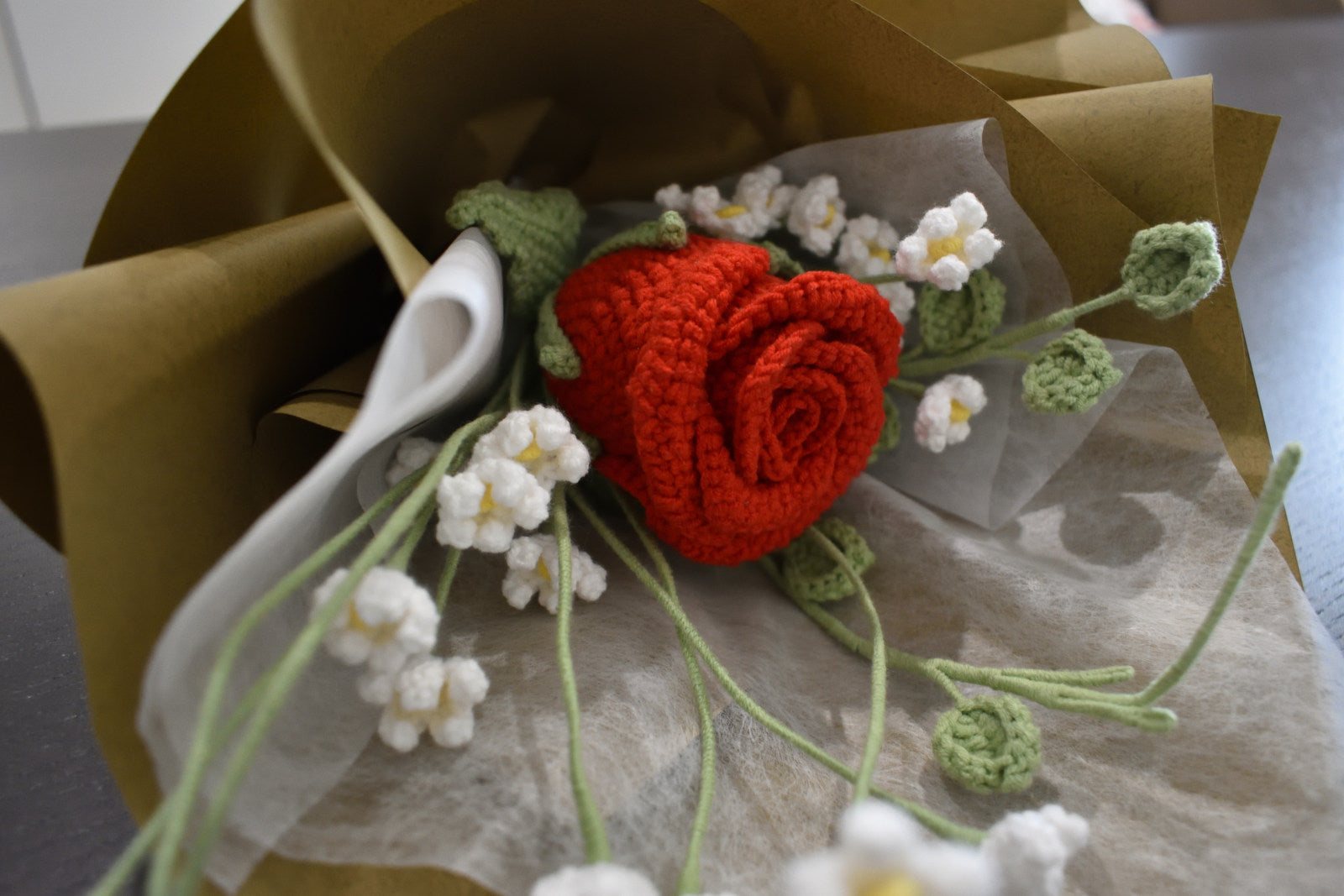 Close-up of a handmade crochet flower bouquet featuring a single red rose with baby's breath and greenery, wrapped in brown paper and laid on a dark table.