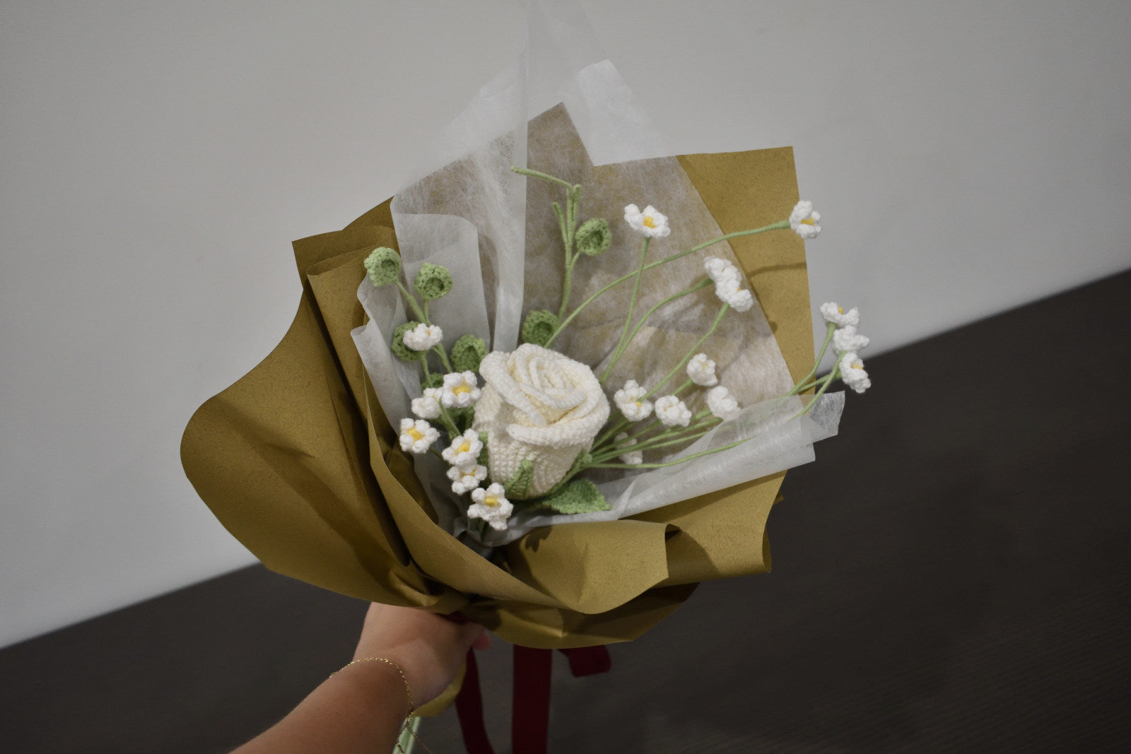 Handmade crochet flower bouquet featuring a single white rose with baby's breath and greenery, wrapped in brown paper and held against a neutral background.
