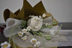 Handmade crochet flower bouquet featuring a single white rose with baby's breath and greenery, wrapped in brown paper and laid on a dark table.
