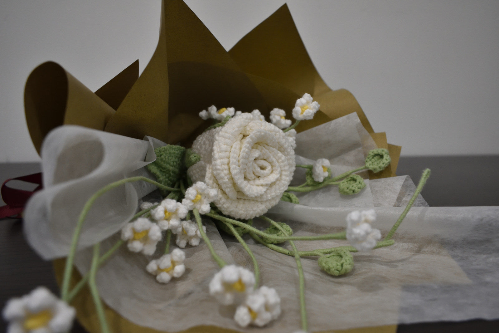 Handmade crochet flower bouquet featuring a single white rose with baby's breath and greenery, wrapped in brown paper and laid on a dark table.