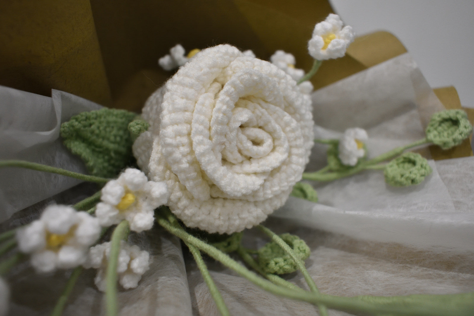 Close-up of a handmade crochet flower bouquet featuring a single white rose with baby's breath and greenery, wrapped in brown paper and laid on a dark table.