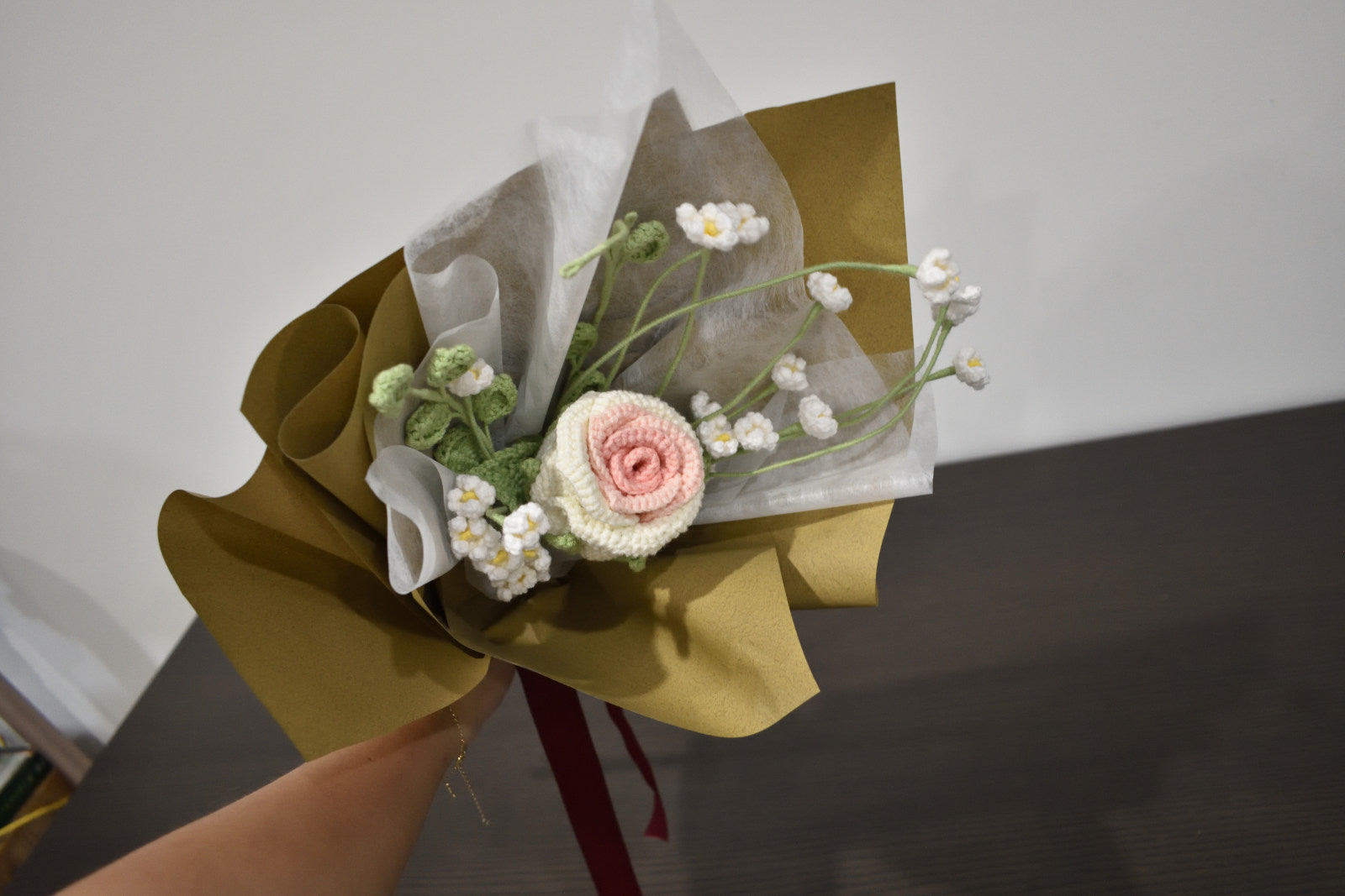 Handmade crochet flower bouquet featuring a single white and pink rose with baby's breath and greenery, wrapped in brown paper and held against a neutral background.