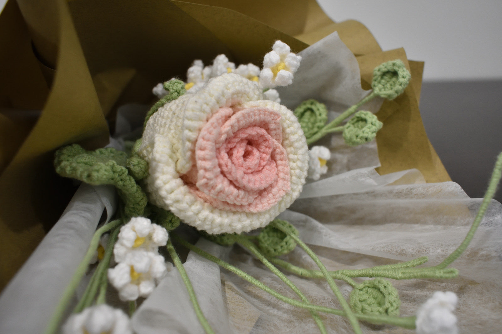 Handmade crochet flower bouquet featuring a single white and pink rose with baby's breath and greenery, wrapped in brown paper and laid on a dark table.