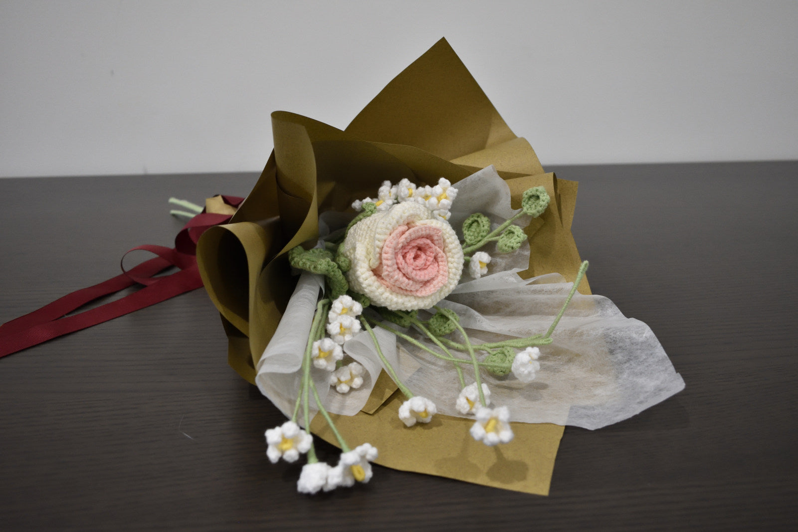 Close-up of a handmade crochet flower bouquet featuring a single white and pink rose with baby's breath and greenery, wrapped in brown paper.