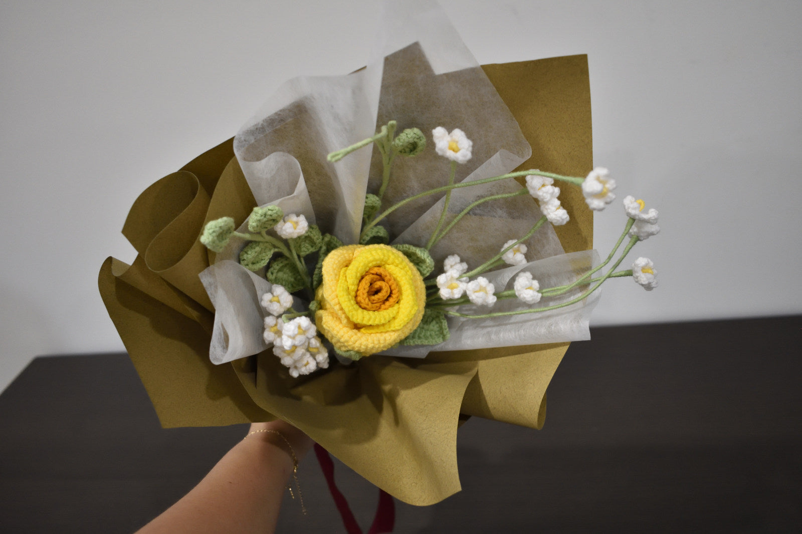 Handmade crochet flower bouquet featuring a single yellow rose with baby's breath and greenery, wrapped in brown paper and held against a neutral background.