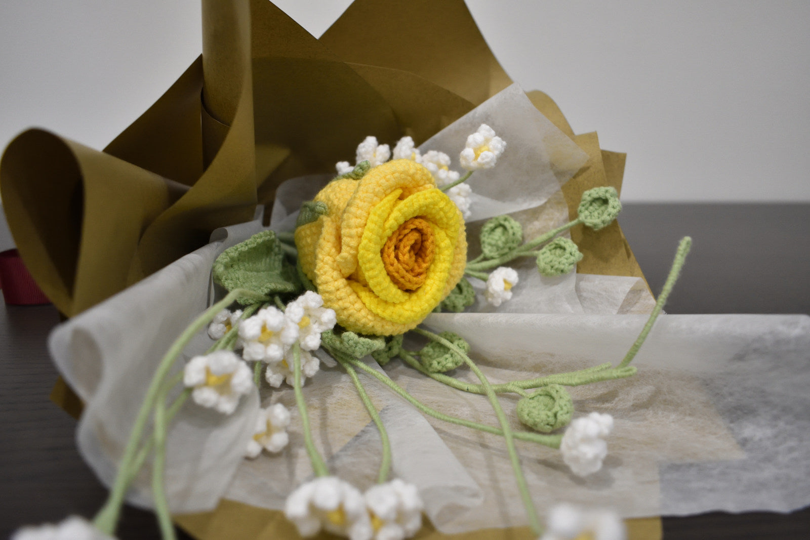 Handmade crochet flower bouquet featuring a single yellow rose with baby's breath and greenery, wrapped in brown paper and laid on a dark table.