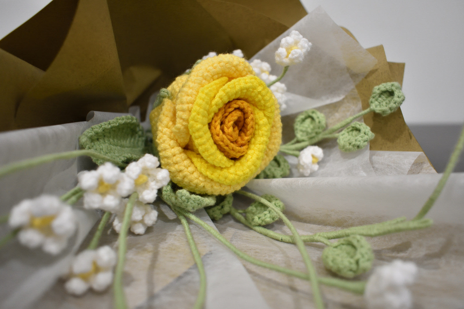 Close-up of a handmade crochet flower bouquet featuring a single yellow rose with baby's breath and greenery, wrapped in brown paper.