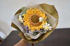 Handmade crochet flower bouquet featuring a single sunflower, with purple and white accent flowers and greenery, wrapped in brown paper and held against a neutral background.
